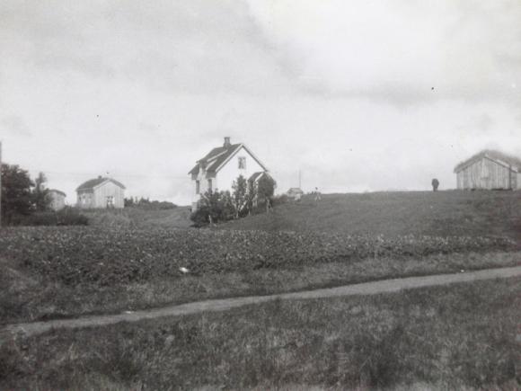 Bakken og &quot;Stasjonen&quot;. �rstallet er ukjent, men muligens f�r 1950 ettersom det st�r en fj�s oppe p� h�yden nord for den siste store fj�sbygningen. Mellom kirkeg�rden og veien en frodig potet�ker. Bildet eies av Judith Hammervold.